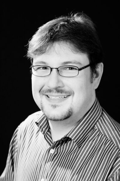 Ein Mann mit Brille und gestreiftem Hemd lächelt vor schwarzem Hintergrund. Schwarz-Weiß-Foto.