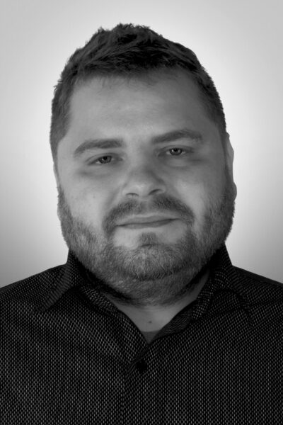 Porträt eines Mannes mit Bart, der in einem gemusterten Hemd vor einem hellen Hintergrund lächelt.